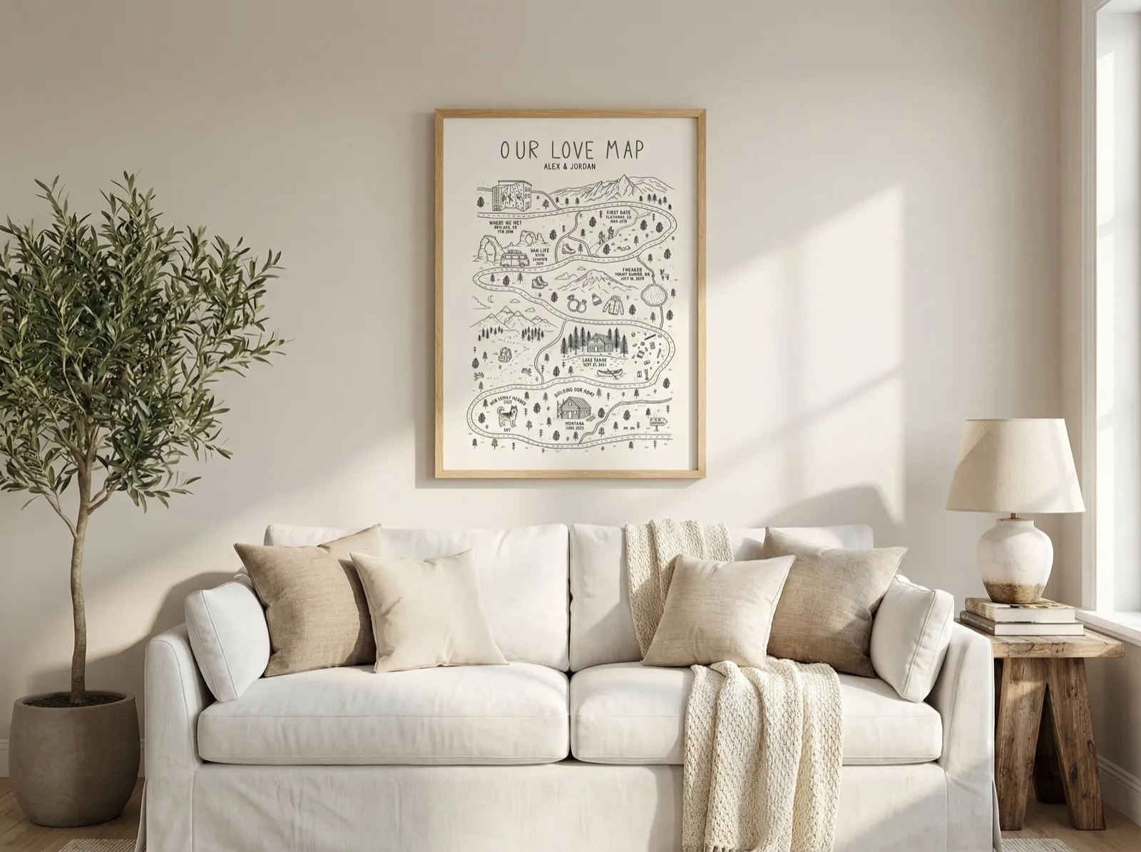 A neutral living room with an olive tree in a pot and a custom love map in a light wood frame hanging above a white sofa with linen cushions.